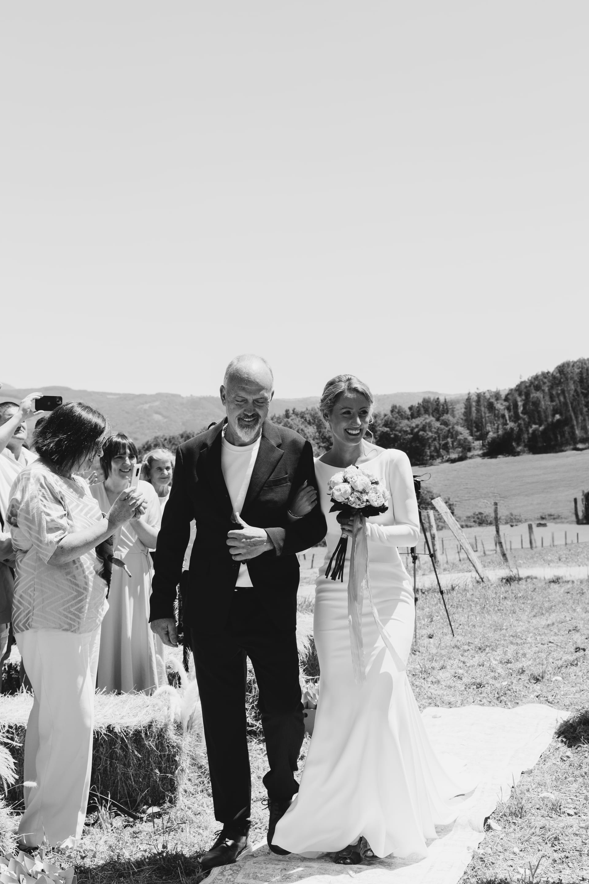 La novia entrando a la ceremonia del brazo de su padre entre los invitados, en blanco y negro